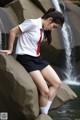 A woman in a school uniform sitting on a rock by a waterfall.